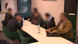 In einem Raum sitzen mehrere Personen an einem Tisch. Die Gesichter der Personen sind verpixelt.