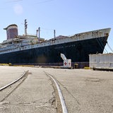 Ein marodes, großes blau-weißes Schiff mit zwei Schornsteinen liegt an einem Pier.