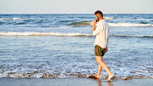 Ein Mann läuft mit einem Hndy am Ohr am Strand entlang.
