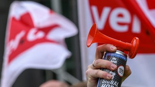 Eine Hand hält eine Fanfare in die Luft, im Hintergrund zwei Verdi-Fahnen