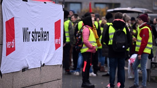 Hinter einem Transparent mit der Aufschrift: "Wir streiken/Verdi", stehen in gelben Warnwesten streikende Menschen.