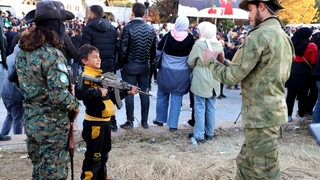 Ein HTS-Kämpfer fotografiert ein kleiner Junge, der gerade seine Kalaschnikow in den Händen hält.