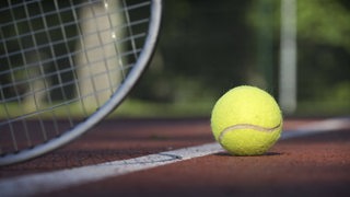 Ein Tennisball liegt auf einem Spielfeld. Im Hintergrund ist ein Teil eines Tennisschlägers zu sehen.