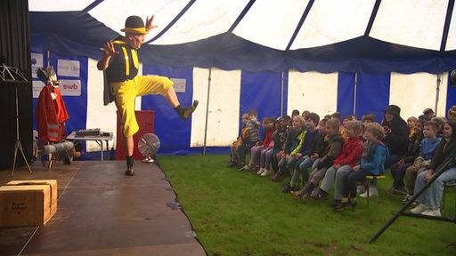 Eine Gruppe Kinder sitzen während einer Theateraufführung auf Bänken in einem Zirkuszelt, ein Mann in gelb steht auf der Bühne.