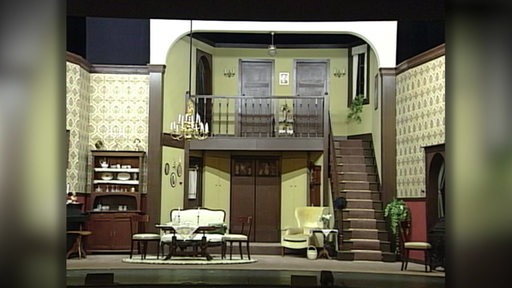 Ein Wohnzimmer ist als Theaterkulisse auf einer Bühne aufgebaut.