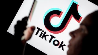 Eine junge Frau im Seitenprofil hält ein Handy hoch mit dem TikTok-Banner im Hintergrund.