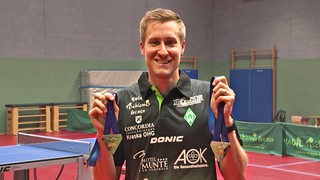 Werder-Tischtennisprofi Mattias Falck hält in der Trainingshalle strahlend seine Gold- und Bronzemedaille der EM in die Kamera.