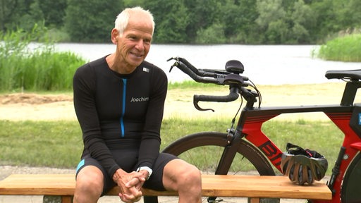 Der Triathlet Joachim Strobach sitzt auf einer Bank vor einem See, neben ihm steht ein rot schwarzes Rennrad.