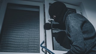 Ein Einbrecher mit Skimaske hebelt ein Fenster mit einer Brechstange auf.