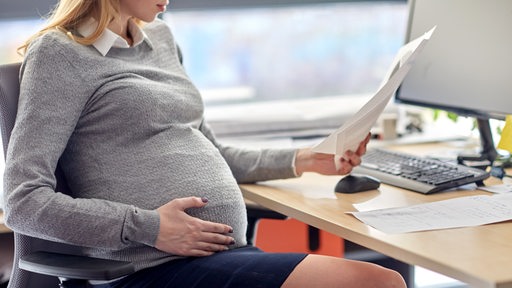 Eine schwangere Frau sitzt in einem Büro und schaut sich Unterlagen an. (Symbolbild)