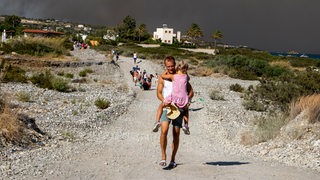 Ein Tourist läuft mit seiner Tochter auf dem Arm aus einem Feriengebiet auf Rhodos. Im Hintergrund rauchschwarzer Himmel