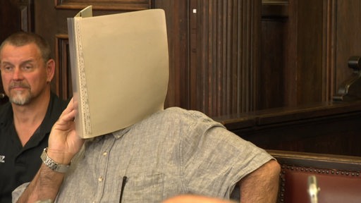 Ein Mann hält sich eine Mappe vor das Gesicht in einem Gerichtssaal.