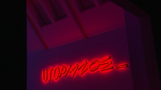 Das rote Leuchtschild einer Kunstausstellung. Das Schild formt den Satz "Utopia now.".