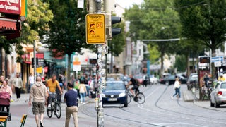 Ein Schild im Bremer Viertel zeigt die Waffenverbotszone an, im Hintergrund ist das Treiben im Viertel zu sehen.