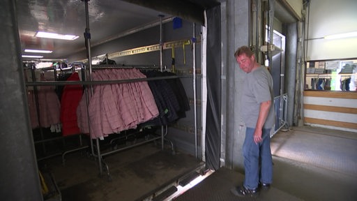 Hinter einem Rolltor ist eine Kleiderstange mit Jacken zu sehen. Vor dem Rolltor steht ein Mann.
