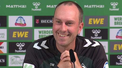 Werder-Trainer Ole Werner lächelt während einer Pressekonferenz ins Publikum.