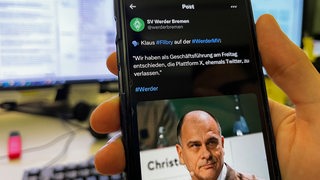 Eine Hand hält ein Handy mit einem Tweet von Werder Bremen im Hintergrund sind PC-Bildschirme zu sehen