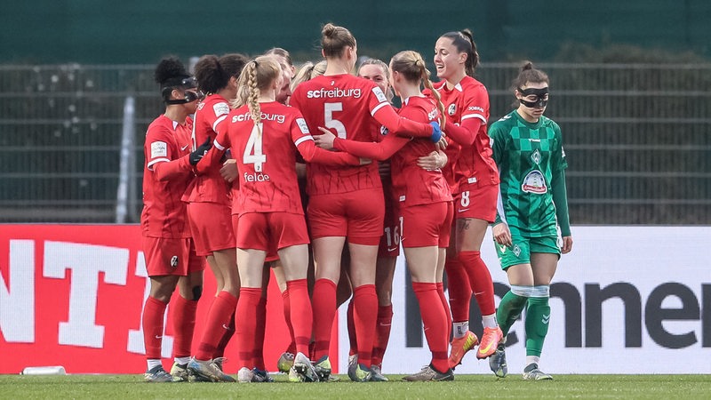 Fußballerinnen in roten Trikots stehen im Kreis und freuen sich, während eine weitere in Grün vorbeigeht.