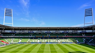 Zu sehen ist das leere Weser-Stadion von innen bei Sonnenschein. Auf der Tribüne steht der Schriftzug "Werder Bremen". 