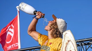 Lennart Neubauer, ein junger Mann, steht auf einem Podium und schüttet sich Champagner in den Mund.