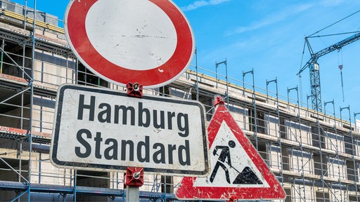 Baustellenschild mit Zusatzschild mit der Aufschrift: "Hamburg Standard" auf Baustelle vor einem Mehrfamilenhaus-Rohbau mit Baugerüsten (Symbolbild)