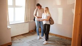 Zwei junge Frauen stehen in einer leeren Wohnung und schauen auf einen Plan.