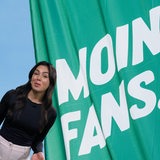 Eine Frau steht vor einem grünen Banner, auf dem in weißer Schrift "Moin Fans" steht.