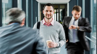 In einem Büroflur sieht man einen ruhigen Angestellten mit Kaffeebecher neben unscharfen Kollegen in Hektik.